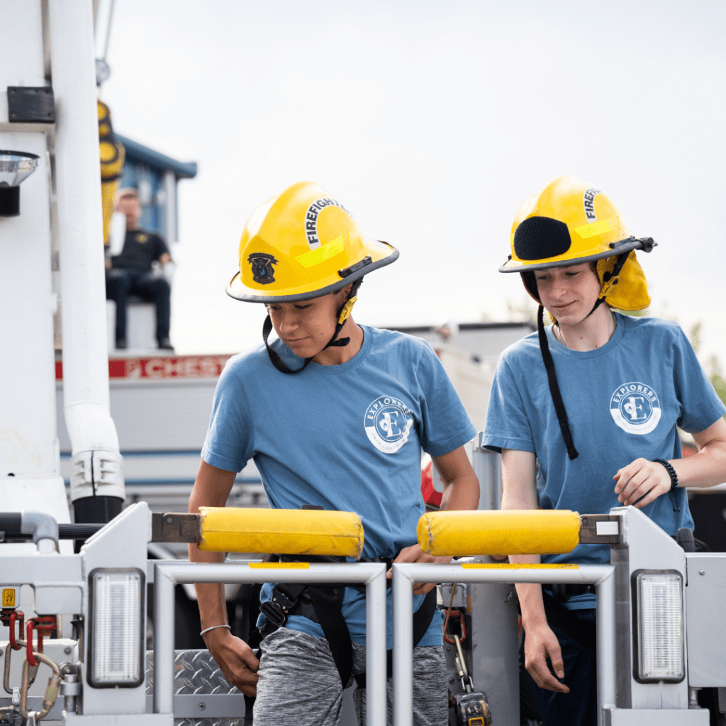 Two young men are seen with firefighter hats on, climbing onto an aerial lift on a firetruck