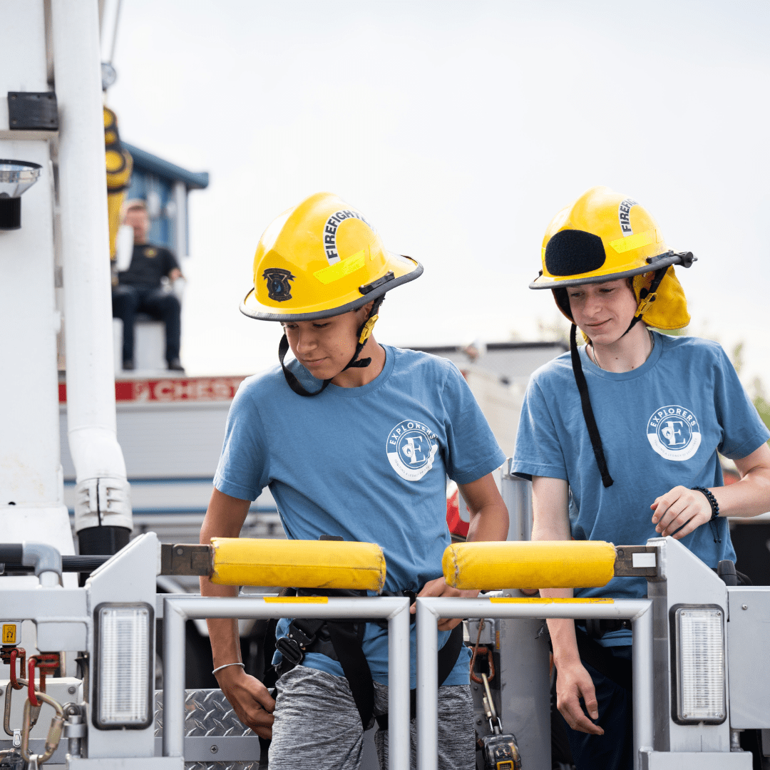 Two young men are seen with firefighter hats on, climbing onto an aerial lift on a firetruck
