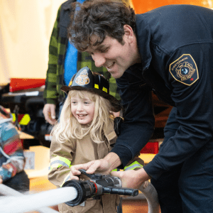 A firefighter is seen assisting a child in operating a hose. They have big smile and appear to be having fun.